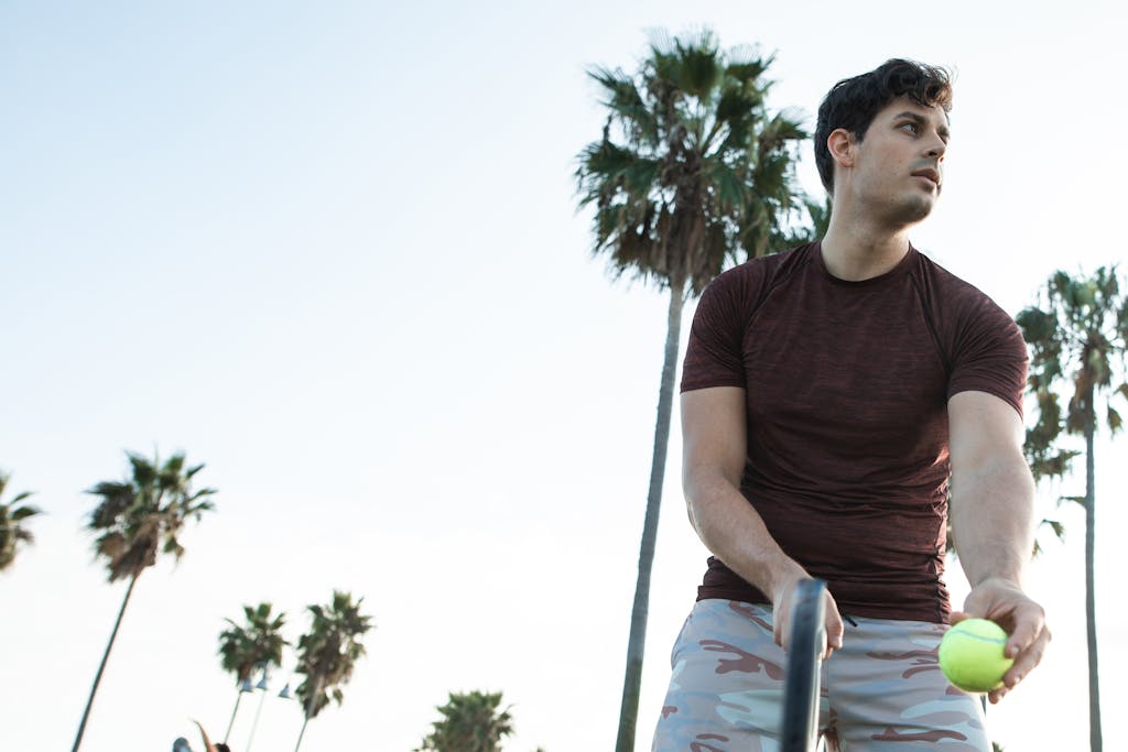 A young caucasian man in casual attire playing tennis outdoors with palm trees in the background.